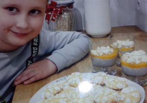 Chłopie siedzący przy stole , na którym widać talerz z gotowymi ciasteczkami i deserem w szklaneczkach.